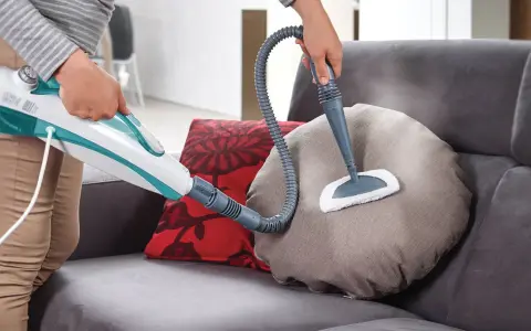Persona pulisce un divano non sfoderabile con vaporetto, igienizzando i cuscini con vapore.
