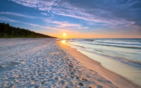 Zachód słońca nad morzem, piaszczysta plaża z ludzkimi śladami, las w tle. Idealne, fajne miejsce nad morzem dla dwojga.