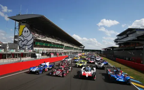 Una fila de coches de carreras listos para comenzar las 24 horas de Le Mans. El icónico muñeco de Michelin preside la tribuna.