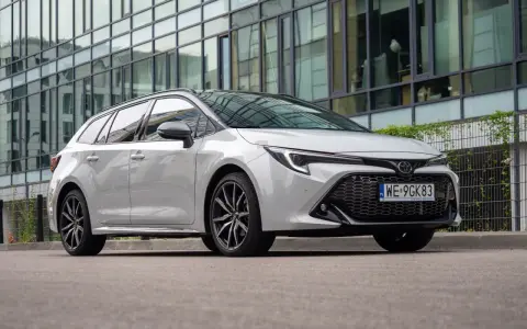 Biała Toyota Corolla 2.0 Hybrid kombi zaparkowana przed nowoczesnym budynkiem z lustrzaną fasadą.