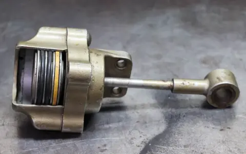 Metalowy element z tłoczyskiem i sprężynami, przypominający zawór wastegate. Objawy uszkodzenia mogą być widoczne na jego powierzchni.