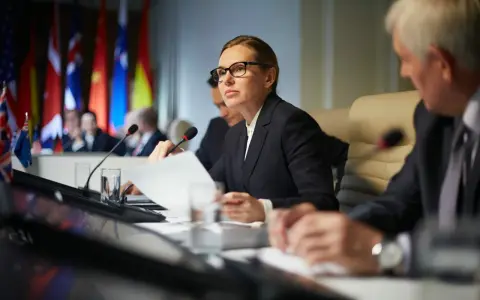 Kobieta w okularach analizuje dokumenty podczas konferencji międzynarodowej, gdzie omawiane są zagadnienia politologii.