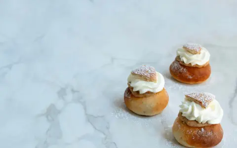 Tre semlor utan mandelmassa, toppade med vispgrädde och pudersocker, ligger på ett marmorbord.