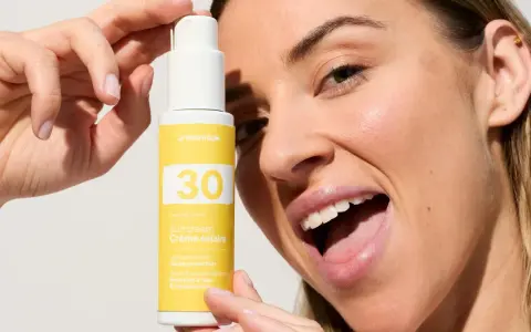 Une femme souriante tient un flacon de crème solaire visage SPF 30. La quantité de crème solaire est parfaite pour une protection optimale.