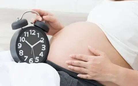 Zbliża się termin porodu. Kobieta trzyma dłoń na brzuchu obok budzika.