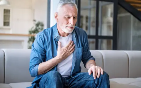 Älterer Mann mit COPD hält sich die Brust. Er sitzt auf einem Sofa und sieht besorgt aus.