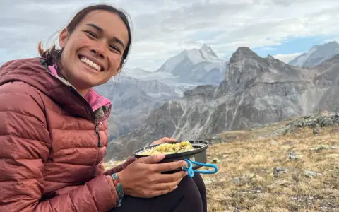 Sourire radieux d'une randonneuse savourant son repas en pleine montagne, avec des sommets enneigés en arrière-plan. Un moment de pur bonheur pour ce repas randonnée.