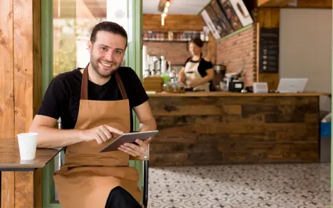 Hombre sonriente con delantal usa tablet en su cafetería, gestionando pedidos con un sistema erp restaurantes.