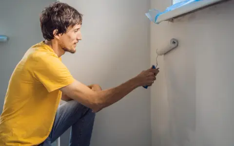 Mężczyzna w żółtej koszulce maluje ścianę wałkiem. Czy przed każdym malowaniem trzeba gruntować ściany? To pytanie zadaje sobie wielu majsterkowiczów.