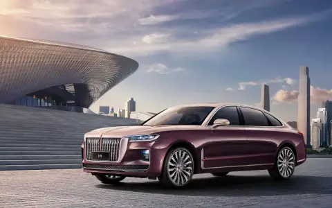 Elegancki, bordowy Hongqi H9 z chromowanymi detalami stoi przed nowoczesnym budynkiem o falistej fasadzie, w tle miejski krajobraz.