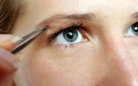 Une main utilise une pince à épiler pour affiner des sourcils, créant une ligne élégante au-dessus d'un œil bleu-vert.