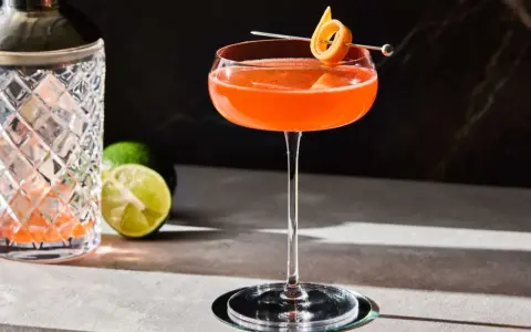 En vackert orangefärgad Division Bell cocktail med en apelsinzest som garnityr, bredvid en cocktailshaker och limeklyftor.