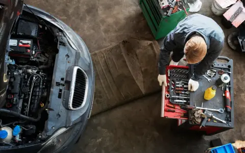 Meccanico con guanti bianchi esamina un motore sbiellato, circondato da attrezzi e un'auto con il cofano aperto.