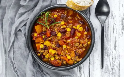 Herzhaftes Chili sin Carne mit Bohnen, Mais und Kürbis, garniert mit Rosmarin. Dazu ein Stück Brot und ein Löffel.