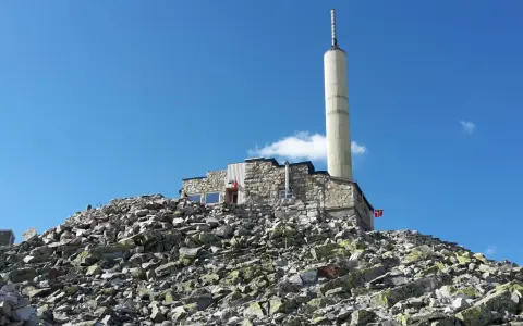 Stone building with a tall tower on a rocky mountain peak under a blue sky.