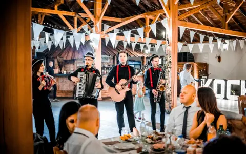 Zespół grający na weselu, z akordeonem, gitarą i saksofonem. W tle widać gości i dekoracje. Idealni do oprawy muzycznej imprez, znają teksty piosenek biesiadnych i weselnych.
