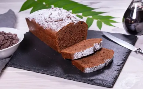 Ein saftiger Schokoladenkuchen, bestäubt mit Puderzucker, bereit für ein köstliches rotweinkuchen rezept. Daneben Schokoladendrops und ein Glas Rotwein.