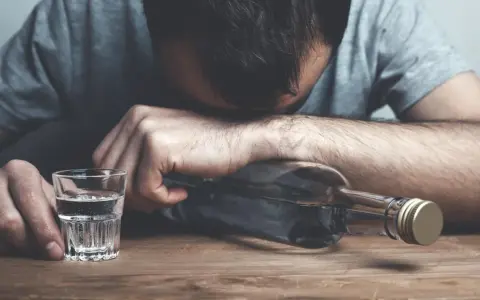 Mężczyzna z głową opartą o butelkę, obok kieliszek z alkoholem. Wygląda na to, że doświadcza zatrucia alkoholowego.