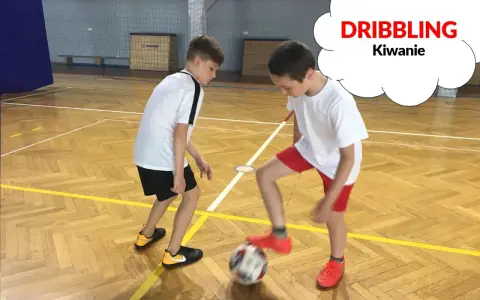 Dwóch chłopców ćwiczy drybling piłką nożną na sali gimnastycznej.