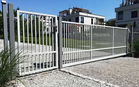 Brama z furtką bez słupka, otwarta na posesję z nowoczesnymi budynkami.
