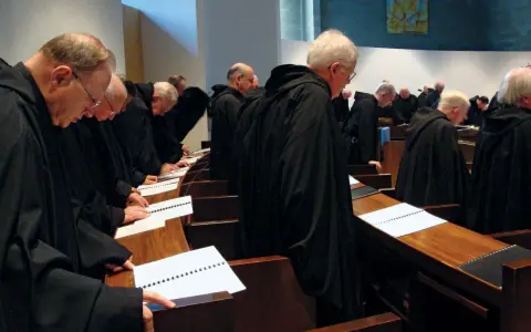 A group of Italian monks in black robes stand in a church, heads bowed over open prayer books.