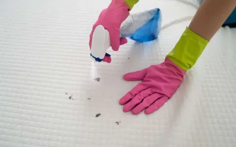 Ręce w różowych rękawiczkach rozpylają środek na plamy na białym materacu, pokazując, jak wyczyścić materac.