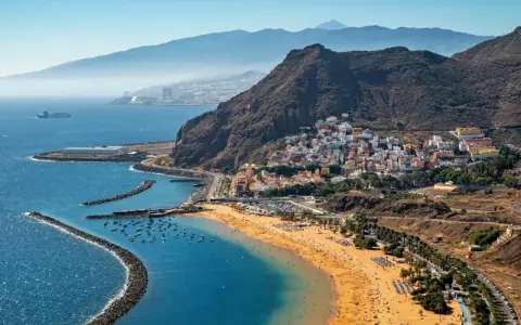 Bunte Häuser schmiegen sich an steile Hänge, während ein goldener Strand und das blaue Meer die Küste von Spanien zieren. Im Hintergrund erheben sich majestätische Berge.