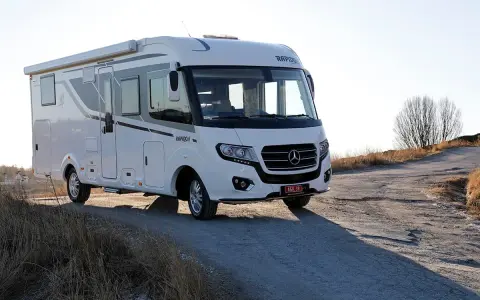 Vit husbil, en Rapido M66, parkerad på en grusväg med torrt gräs och träd i bakgrunden.