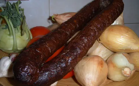 Wędzona kiełbasa z dzika na talerzu z warzywami: cebulą, pomidorami, kalarepą i czosnkiem.