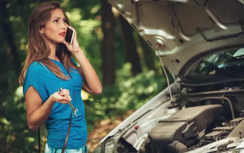 Mujer llama por teléfono con una llave inglesa en la mano, preguntando por qué se me para el coche en marcha. El capó del coche está abierto.