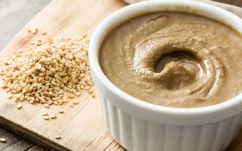Un bol de sauce tahini crémeuse, à côté d'une pile de graines de sésame sur une planche en bois.
