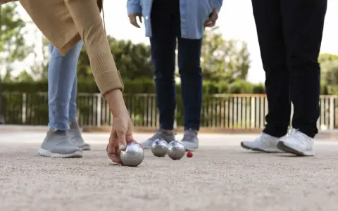 Une main de nom joueur de pétanque français saisit une boule argentée sur un terrain de gravier. D'autres boules et un cochonnet sont visibles.
