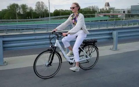 Kobieta w białych spodniach i sweterku jedzie na rowerze elektrycznym. W tle widać most i budynki.