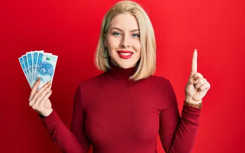 Kobieta w czerwonym golfie trzyma polskie banknoty i pokazuje palcem do góry.