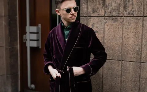 Elegancki mężczyzna w bordowej bonżurce z aksamitu, w okularach przeciwsłonecznych, stoi przy kamiennej ścianie.
