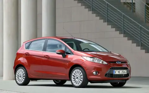 Czerwony Ford Fiesta MK6 zaparkowany przed nowoczesnym budynkiem z betonowymi kolumnami i schodami.
