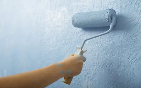 Mano aplica imprimación azul con rodillo a pared texturizada. La imprimación es el primer paso para un acabado perfecto.