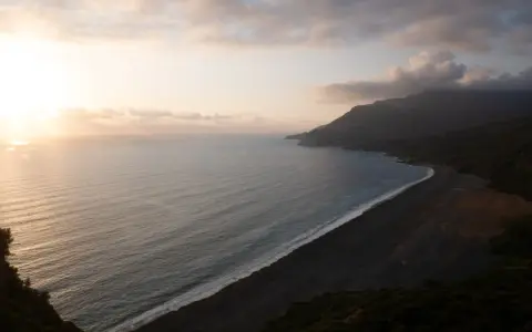 Vue aérienne d'une plage de sable noir bordée par une mer calme et des montagnes embrumées. Ces photos de plage capturent la sérénité d'un paysage côtier au coucher du soleil.