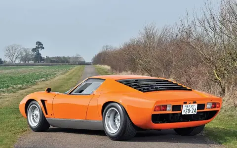 Ein leuchtend oranger Lamborghini Jota parkt auf einer einsamen Landstraße, umgeben von Feldern und kahlen Bäumen.