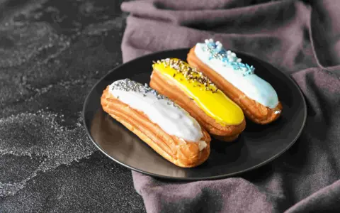 Trois éclairs gourmands sur une assiette noire : un blanc pailleté, un jaune croquant, un bleu ciel festif. La pâte à choux éclair inratable est prête à être dégustée.