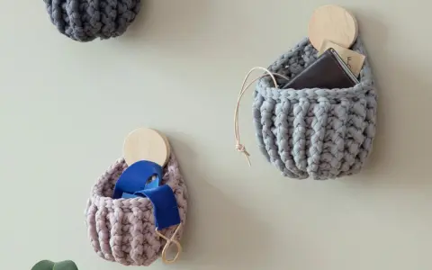 Three crocheted wall pockets in grey and pink, one holding a wallet, another blue ribbon, on a light wall.