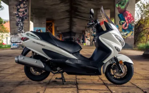 Biały skuter Suzuki Burgman 125, z czarnym siedzeniem, zaparkowany pod mostem z kolorowymi graffiti. Idealny do miasta, sprawdź suzuki burgman 125 dane techniczne.