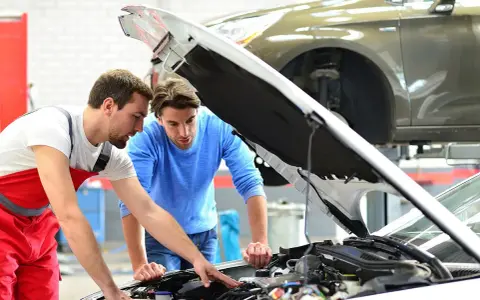 Zwei Männer blicken in den offenen Motorraum eines Autos. Der Mechaniker in Rot erklärt dem Kunden, welche Werkstatt mitgebrachte Teile einbaut.