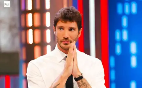 Santiago de Martino, con le mani giunte in preghiera, guarda intensamente verso l'obiettivo, con uno sfondo di luci colorate.