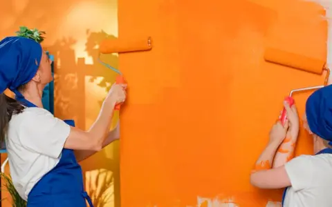 Dos personas pintan una pared de naranja brillante con rodillos. La pintura mural exterior cobra vida con vibrantes tonos anaranjados.