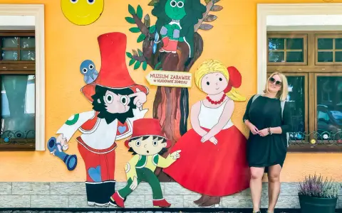 Kobieta pozuje przy kolorowej ścianie Muzeum Zabawek w Kudowie-Zdroju, Dolny Śląsk. Wesołe postacie z bajek towarzyszą jej w radosnej scenerii.