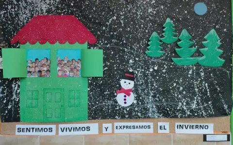 Decoración invierno aula infantil: una casa verde con fotos de niños, árboles, muñeco de nieve y luna sobre fondo negro salpicado.