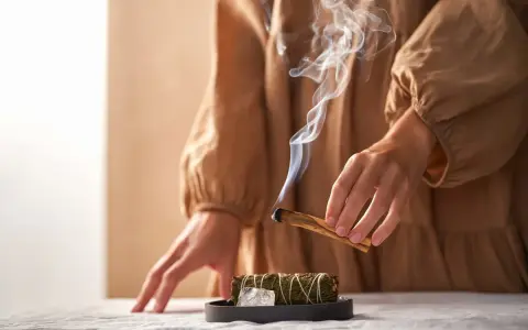 Dym z kadzidła unosi się, tworząc mistyczną atmosferę. Ręce trzymają drewno, symbolizując rytuały związane z magią i okultyzmem.