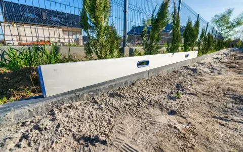 Poziomica na krawężniku, element uzbrojenia działki krok po kroku. W tle ogrodzenie i tuje.