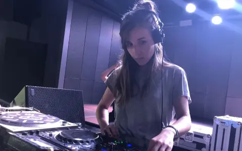 Une dj fille concentrée aux platines, casque sur les oreilles, dans une ambiance tamisée avec des lumières bleues.